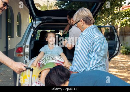 Famiglia caucasica che va in vacanza di viaggio su strada, il caricamento di bagagli e valigia in bagagliaio auto. Genitori e nonni partono in vacanza con la bambina, in viaggio verso il mare. Foto Stock