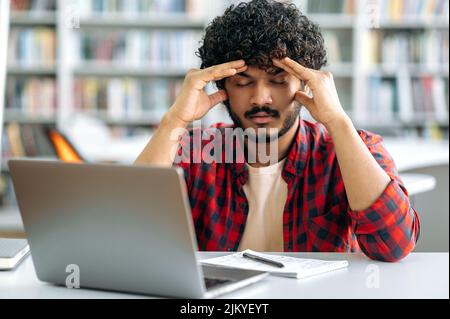 Stanco ragazzo indiano o arabo teso, studente universitario, indossando una camicia elegante, si siede a un tavolo in biblioteca, chiuso gli occhi, massaggi il suo whiskey, stanco di noiosi studi, ha un mal di testa, ha bisogno di rottura Foto Stock