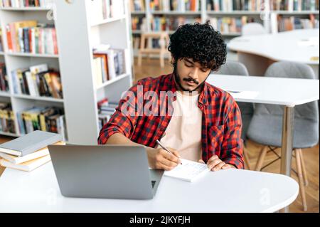 Accecato studente arabo o indiano, si siede al tavolo con il portatile in biblioteca universitaria, preparazione per l'esame, studio di informazioni, ascolto di lezioni, prendere appunti in notebook, acquisire conoscenze Foto Stock