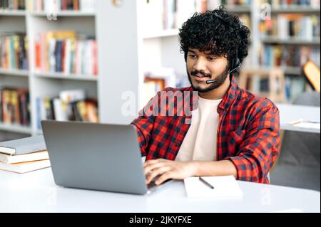 Studente universitario maschile soddisfatto, di nazionalità araba o indiana, con cuffie, siede a un tavolo con un laptop in biblioteca, studia online, ascolta una lezione, webinar, si prepara per un esame Foto Stock