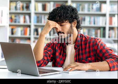 Esausto studente indiano triste o arabo maschio universitario, in una camicia elegante, si siede a un tavolo in biblioteca, chiuso gli occhi, massaggi naso ponte, stanco di noiosi studi, ha un emicrania, ha bisogno di rottura Foto Stock