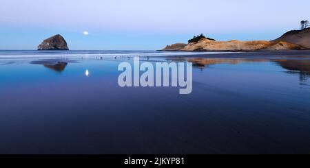 Spiaggia panoramica a Pacific City Oregon con haystack rock e Cape Kiwanda promontorio in luce all'alba come luna piena tramonta Foto Stock