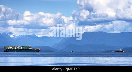 Una chiatta contenitore molto piena che viene tirata da un grande rimorchiatore giù lo stretto con uno sfondo di montagne e grandi nuvole soffici. Foto Stock