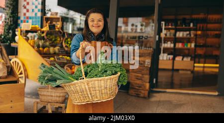 Dipendente del negozio con sindrome di Down che tiene un cestino di verdure fresche biologiche in un negozio di alimentari. Donna felice con una disabilità intellettuale che lavora Foto Stock