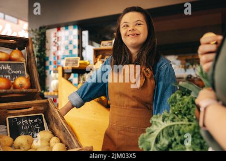 Donna felice con sindrome di Down assistere un cliente nella sezione di verdura di un negozio di alimentari. Donna amichevole con un lavoro di disabilità intellettuale Foto Stock