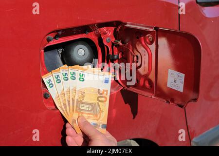 Mano che tiene un pacchetto di cinquanta banconote di euro davanti ad un serbatoio di carburante, aumento del prezzo della benzina. Foto Stock