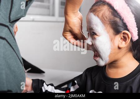 Madre asiatica e figlia capretto ottenere trucco halloween faccia pittura assomiglia a fantasma, ritratto della donna che applica la spazzola di colore alla faccia della ragazza del bambino per pa Foto Stock