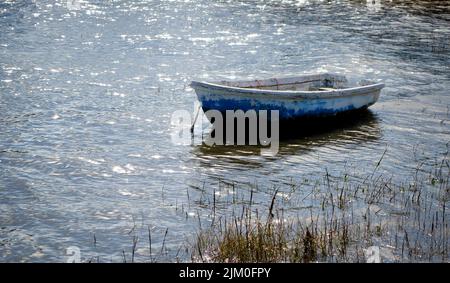 Una piccola barca a remi blu siede su un mare scintillante illuminato dal sole che trasmette un senso di calma e tranquillità. Foto Stock
