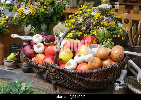 Cestino pieno di mele, cipolle, aglio e carciofi, raccolto dall'orto decorato con mazzi di fiori selvatici su un rustico tovagliolo di legno Foto Stock