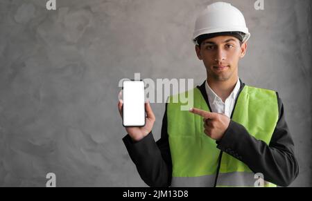 Uomo arabo sorridente ingegnere o costruttore in casco bianco e mostra smartphone con schermo vuoto su sfondo grigio. Professione Foto Stock
