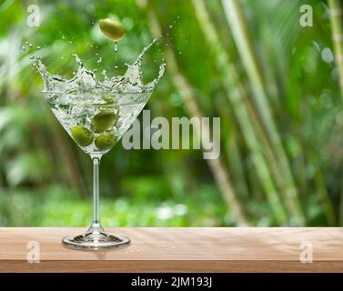 Bicchiere Martini con olive e bevande splash è sul tavolo. Natura sfocata verde sullo sfondo. Inserire il nome del prodotto o della marca. Foto Stock