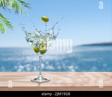 Bicchiere Martini con olive e bevande splash è sul tavolo. Mare frizzante sfocato sullo sfondo. Inserire il nome del prodotto o della marca. Foto Stock