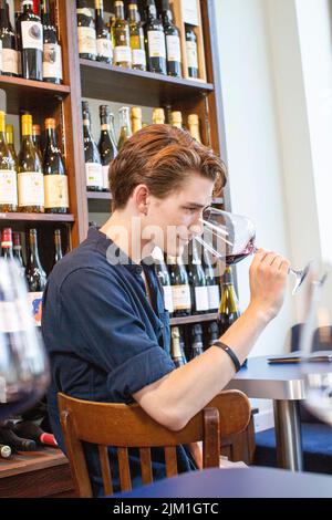 Giovane maschio odore sapore di vino rosso in ristorante sullo sfondo di mensole con bottiglie. Uomo degustazione un bicchiere di vino rosso. Foto Stock