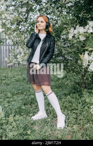 Una ragazza adolescente dai capelli rossi ascolta la musica in cuffie, configurate tramite telefono. Il tempo della fioritura primaverile di ciliegi Foto Stock