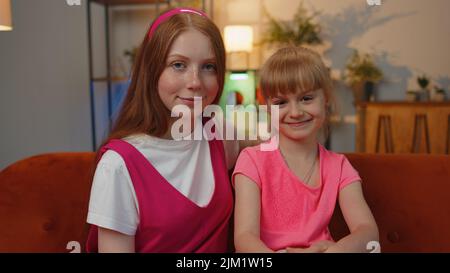 Primo piano ritratto di felice sorridente due ragazze adolescenti amici fratelli guardando la macchina fotografica seduta sul divano di casa. Femmina positivo bambino bambini famiglia coppia winking, occhi lampeggianti. Amicizia fidanzate Foto Stock