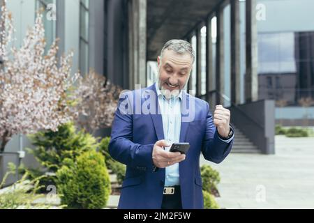 Ritratto di successo senior grigio-capelli banchiere fuori ufficio edificio, uomo con telefono sorridente e gioire in vittoria, festeggiare trionfo, leggere buone notizie dal telefono cellulare Foto Stock