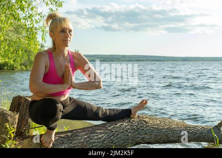 Donna anziana in abiti sportivi che praticano lo yoga in piedi a metà passo e tenendo le mani ripiegate namaste su un albero sulla riva del lago. Foto Stock