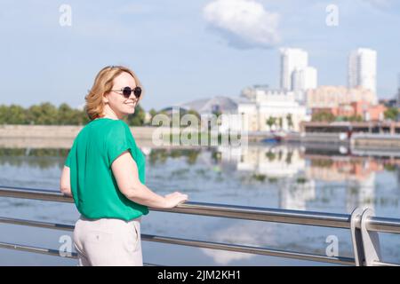 Una vista dal retro di una ragazza in una blusa verde in piedi sul fiume sullo sfondo di una città sfocata. Una ragazza cammina per la strada Foto Stock