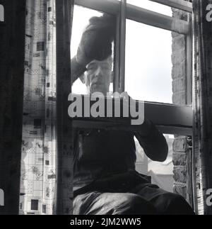 1960s, storico, un uomo che si stende da una finestra a telaio di legno, pulendo il vetro, Inghilterra, Regno Unito. Foto Stock
