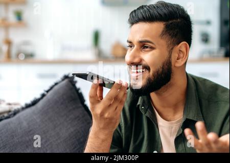Giovane indiano o arabo positivo in abbigliamento casual, sedersi su un divano in un accogliente soggiorno, tenere il suo smartphone, parlare con vivavoce e sorridere, registrare messaggio vocale. Concetto di tecnologia wireless Foto Stock