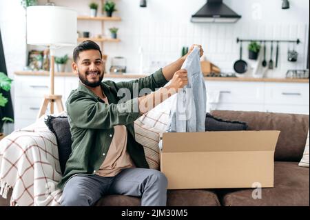 Concetto di shopping online. Gioioso arabo o indiano giovane uomo disimballaggio il suo pacco, felice di ottenere un lungo atteso ordine. Misto razza moderno ragazzo shopping in negozi internet, l'acquisto di nuovi vestiti online Foto Stock