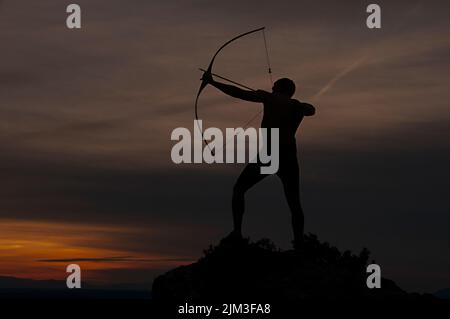 Silhouette di un bell'uomo con un'antica arma a prua e freccia su uno sfondo di cielo e tramonto. Foto Stock