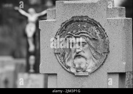 Un focus selettivo del volto scolpito di Gesù Cristo sullo sfondo sfocato di Cristo crocifisso sulla croce nel cimitero di Pecs, Ungheria Foto Stock