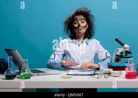 MAD scienziato guardare seduto alla scrivania in laboratorio con capelli disordinati e volto sporco dopo esperimento chimico fallito. Maniaco goofy chemist con aspetto esploso e fumetto guardando la fotocamera. Foto Stock