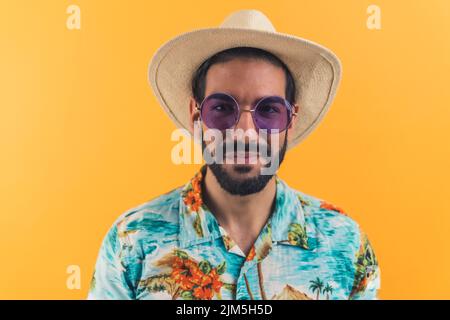 Uomo ispanico con barba che indossa occhiali da sole con cappello di paglia e camicia a manica corta modellata che guarda nella macchina fotografica. Turistico. Foto studio. Foto di alta qualità Foto Stock