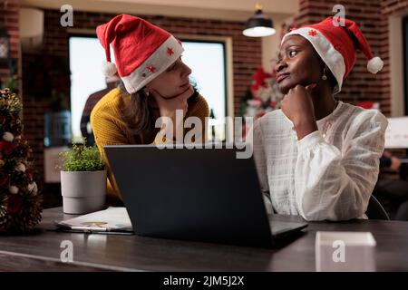 Team diversificato di colleghi con babbo Natale che lavorano per il business al computer portatile, seduti in ufficio festivo pieno di decorazioni e ornamenti di natale. Fare lavoro di squadra e celebrare le vacanze. Foto Stock