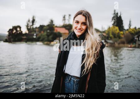 Giovane donna bionda calda in piedi e sorridente di fronte ad un bel lago in un pomeriggio nuvoloso. Spazio di copia Foto Stock