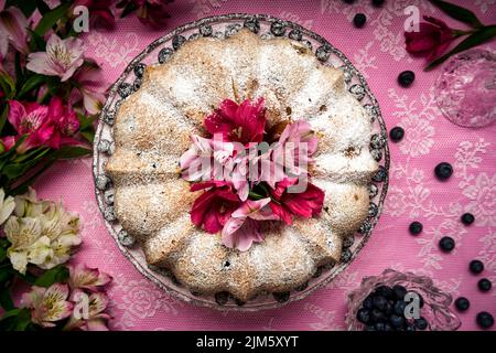 Una vista dall'alto di una torta di mirtilli al limone con zucchero a velo e decorata con fiori su sfondo tovaglia in pizzo rosa Foto Stock