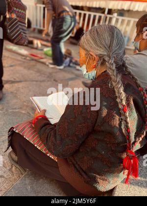 Un'immagine di una vecchia donna con trecce seduta a terra e leggendo un libro in Tibet, Cina Foto Stock