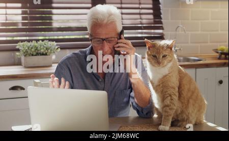 I miei clienti sanno che sono sempre pronti ad aiutarli. Un uomo anziano seduto da solo in cucina con il suo gatto e utilizzando la tecnologia per lavorare da casa. Foto Stock