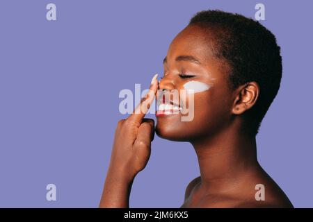 Afro donna applicazione crema sulle sue guance e con il suo indice dito diffonde la crema sul suo naso isolato su sfondo viola con spazio vuoto per Foto Stock