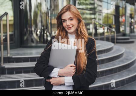 Ritratto all'aperto di una donna zenzero che tiene un computer portatile e sorride alla fotocamera mentre si va a casa dopo il lavoro. Concetto di lavoro remoto. Donna d'affari di successo Foto Stock