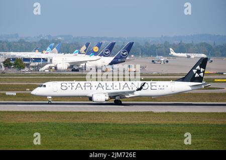 Un aereo Star Alliance decollerà di fronte ad altri aerei parcheggiati al terminal dell'aeroporto di Monaco, con partenza da Embraer 190. Foto Stock