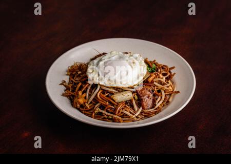 Un primo piano di una spaghetta fritta con uovo laterale soleggiato in un piatto bianco su un tavolo di legno Foto Stock