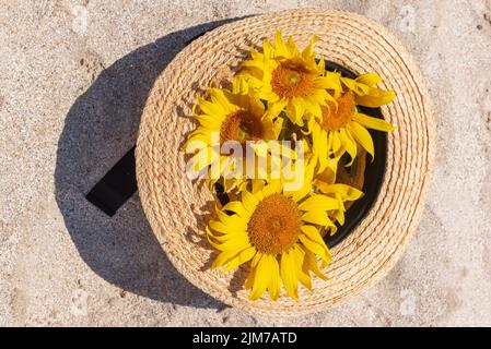 Vista dall'alto di un cappello di paglia pieno di girasoli, adagiato sulla sabbia del mare, da vicino. Concetto di relax e libertà. Foto Stock