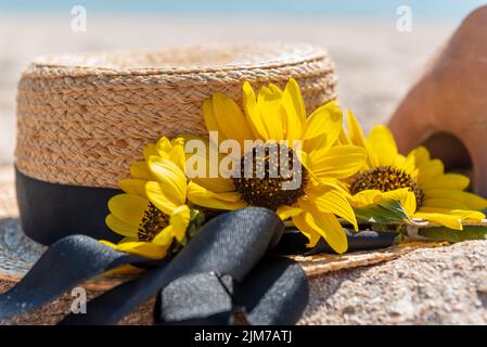 Boater con nastro nero, decorato con girasoli, adagiato sulla sabbia su sfondo mare sfocato e frammento di tacchi alti da donna in piedi nelle vicinanze. Foto Stock