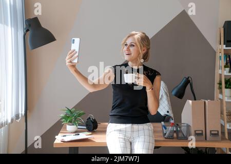 Condividi buone notizie aziendali. Giovane donna attraente che parla al telefono cellulare tramite collegamento video e sorridente, sul suo posto di lavoro in ufficio in una blusa nera e pantaloni bianchi Foto Stock