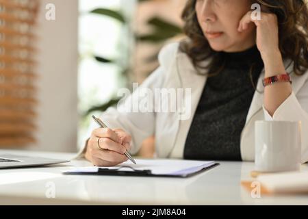 Donna matura focalizzata sul lavoro a distanza da casa, scrivendo informazioni sul notebook Foto Stock
