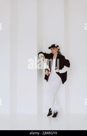 Una giovane donna elegante in un abito alla moda, un abito bianco chiaro con piume, una giacca nera, un cappello da baseball e un bouquet di fiori in mano Foto Stock