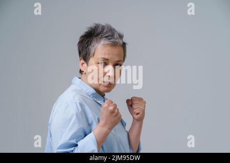 Donna spaventata con le mani messe in pugni. Donna anziana con capelli grigi posa come pugilato combattente circa per stare in piedi o proteggersi. Concetto di tratta di esseri umani. Donna sicura, forte e aggressiva. Foto Stock