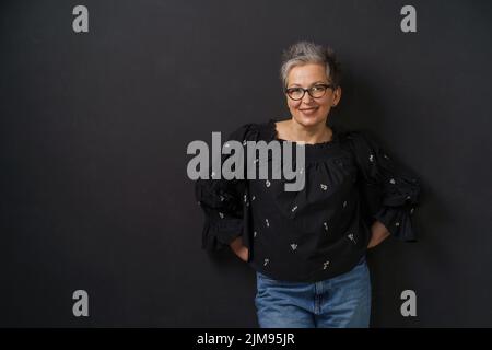 Splendida donna d'affari grigia matura con capelli grigi appoggiata su parete isolata su sfondo nero. Donna elegante con capelli corti. Immagine di una donna matura di successo. Bellezza invecchiata. Foto Stock