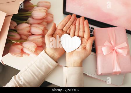 Le mani della donna che tengono carta cuore scheda. Sfondo rosa, concetto di San Valentino. Tulipani fiore e scatola regalo rosa in background. Scrivania per donne. Foto Stock