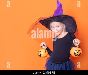 Una ragazza in costume da strega in un cappello che tiene una lanterna di zucca jack con dolcetti su sfondo arancione. Il concetto di Halloween. Festa di Halloween. Foto Stock