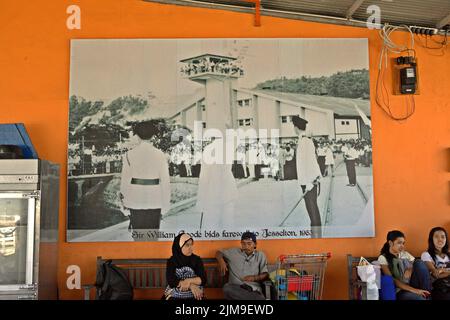 I passeggeri in attesa del trasporto sono seduti davanti a un muro dove una grande stampa fotografica di un momento storico è appesa a Jesselton Point, un porto per le barche che collegano le isole adiacenti con Kota Kinabalu, che si trova nella capitale dello stato di Sabah a Sabah, Malesia. Foto Stock