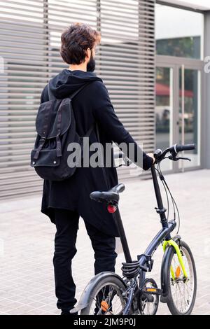 vista posteriore di un giovane uomo che indossa abiti alla moda e cammina con la sua bicicletta pieghevole in città, concetto di stile di vita urbano e mobilità sostenibile Foto Stock