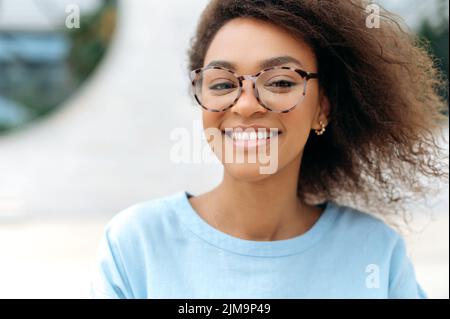 Primo piano di una bella carismatica giovane donna afroamericana con capelli ricci, indossare occhiali, anche con denti bianchi, stare in piedi all'aperto, guardare la macchina fotografica, sorridente amichevole Foto Stock